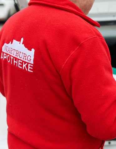 Mitarbeiter der Wartburg Apotheke trägt einen roten Fleece mit dem Logo auf dem Rücken. In der Wartburg Apotheke in Eisenach